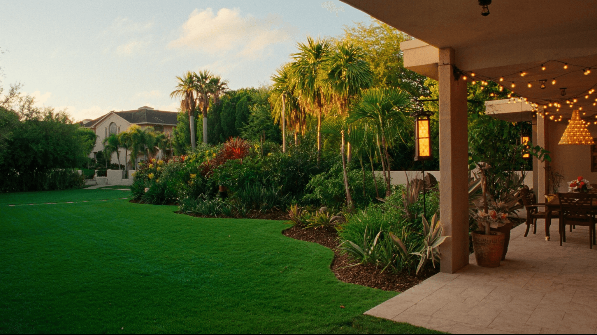 A finished tropical backyard at golden hour — lush green lawn bordered by layered planting beds and palms, warm light raking across the grass